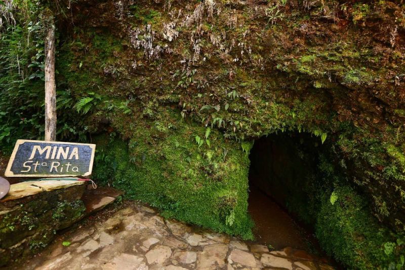 Entrada da Mina Santa Rita em Ouro Preto, com parede de pedra coberta por vegetação verde e placa de madeira indicando o nome da mina